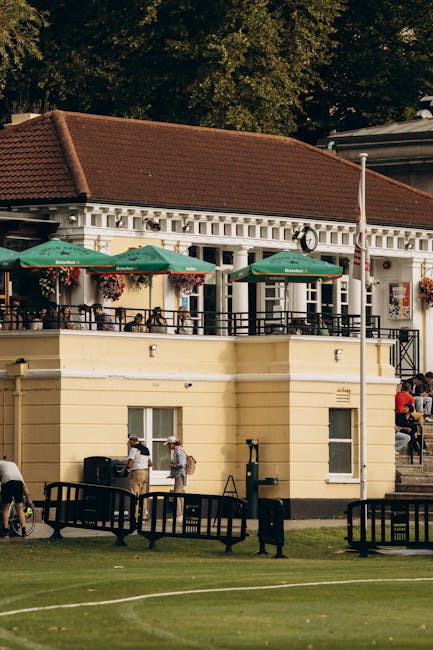 The image depicts a two-story building with a classic design, featuring light yellow walls and a red-tiled roof. The upper level has a balcony with black railings, decorated with hanging flower basket
