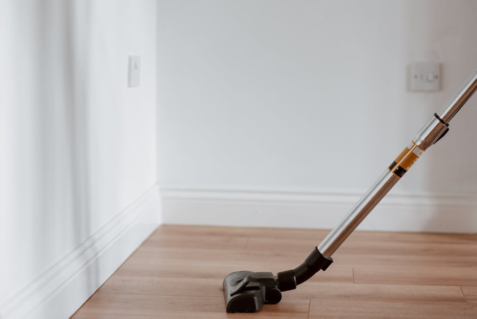 A woman with long blonde hair wearing a dark green T-shirt is holding a clipboard and writing with a black marker. She is standing in a room with a light-colored wall and wooden flooring. Behind her, 