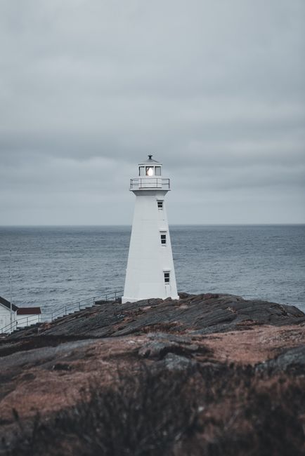 The image features a white lighthouse situated on a rocky shoreline with dark, textured rocks in the foreground. The lighthouse stands tall against a cloudy, overcast sky, with a metal observation dec